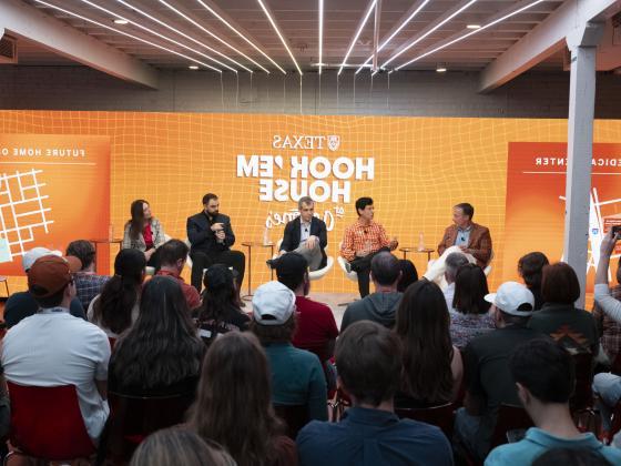 group of people on a panel in front of a crowd in chairswith a burnt orange hook 'em house logo in the background on a screen